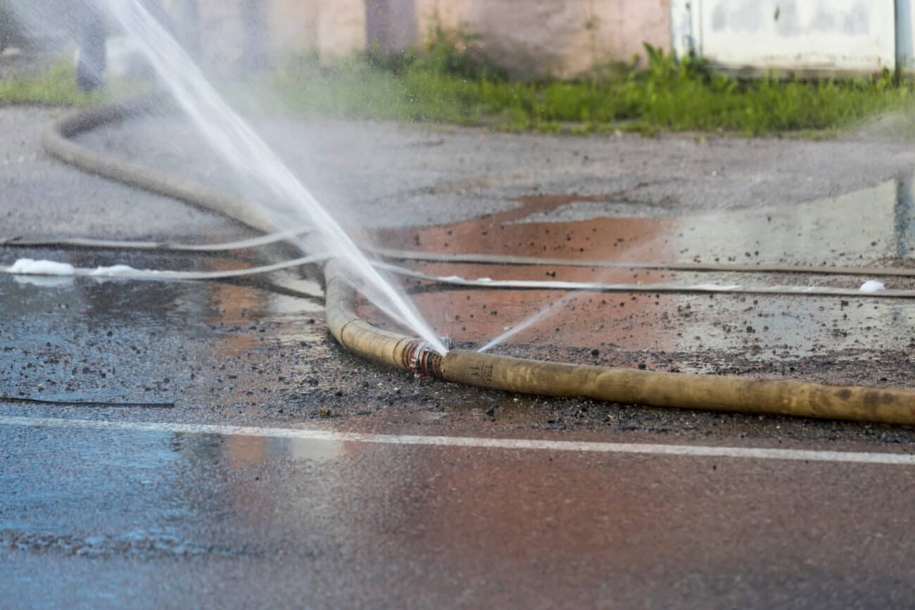 Water sprays aggressively from a fire hose onto the paved street, creating a refreshing mist on a warm afternoon in the city.